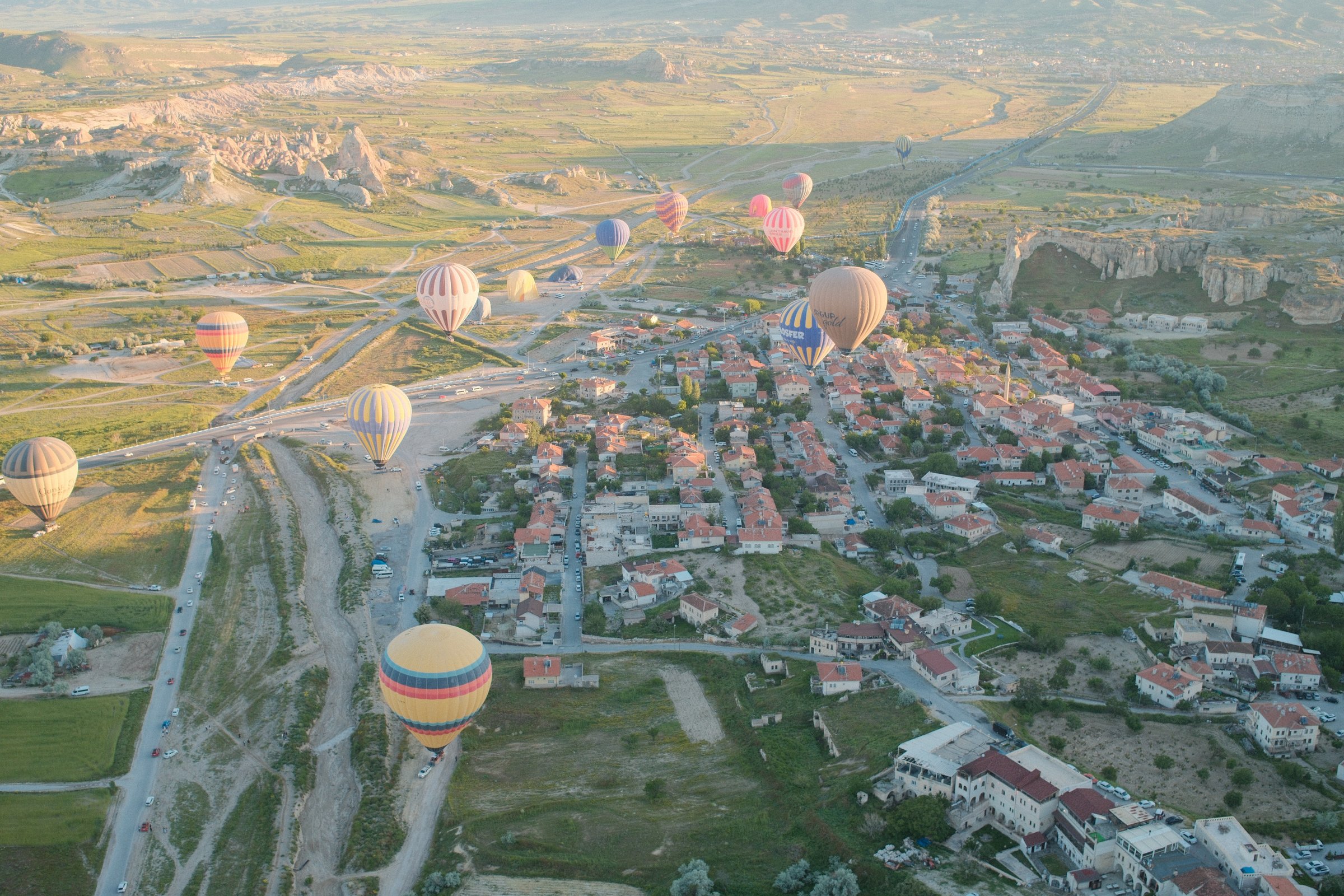 Dozens of balloons descending over a village at sunrise, rock formations visible in the distance.