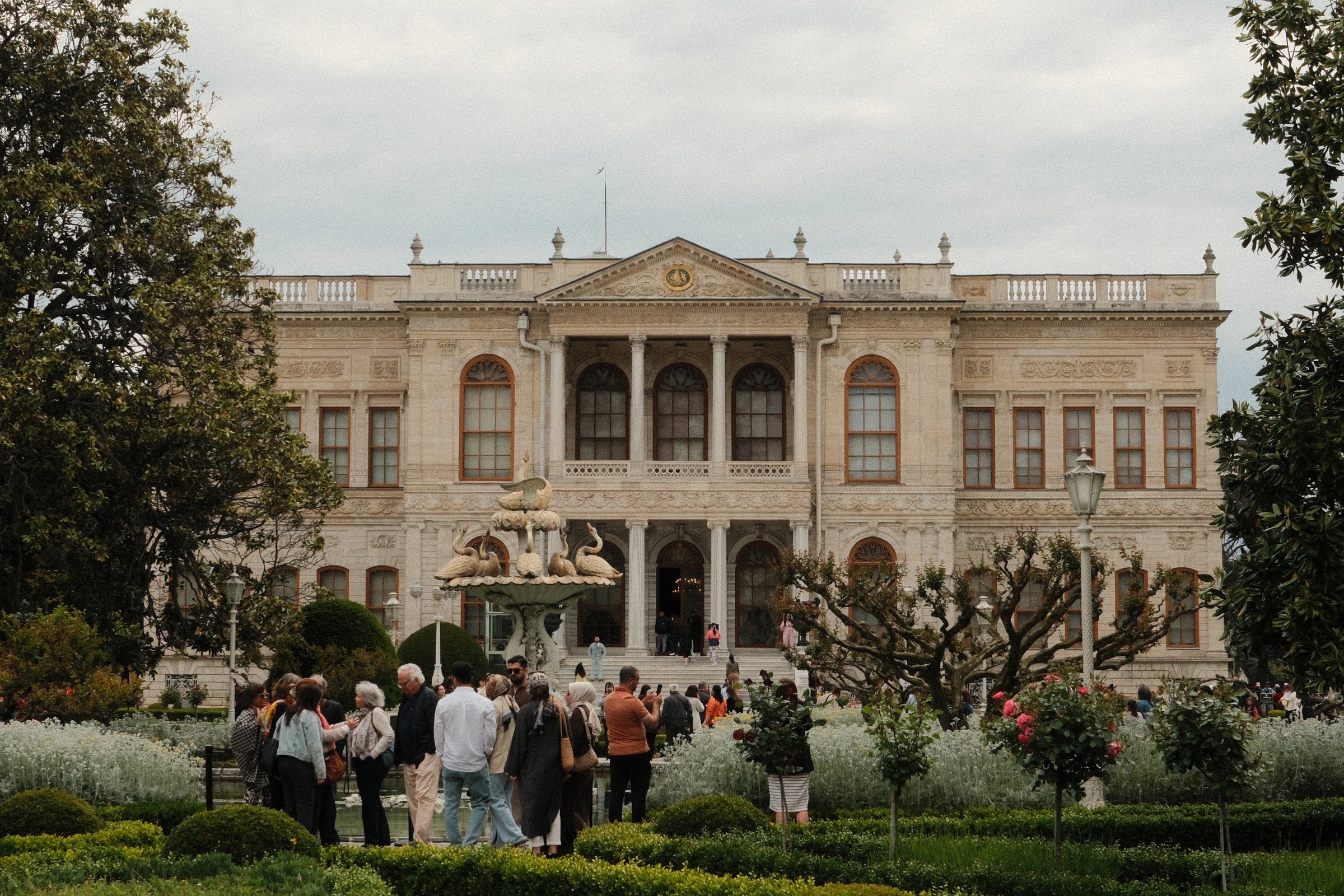Dolmabahce Palace's grand facade behind a swan fountain, visitors gathered in the gardens out front.